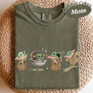 May include: Moss green t-shirt featuring a cartoon character from a popular science fiction franchise. The character is shown in different poses, with a frog, butterflies, and musical notes. The shirt is labelled "Comfort Colors".
