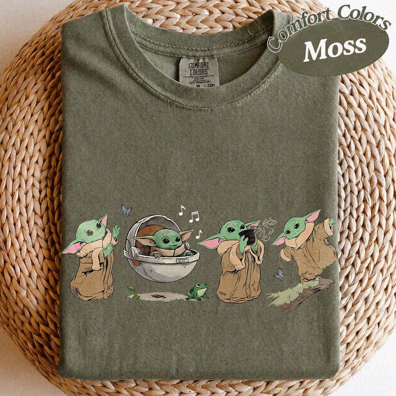 May include: Moss green t-shirt featuring a cartoon character from a popular science fiction franchise. The character is shown in different poses, with a frog, butterflies, and musical notes. The shirt is labelled "Comfort Colors".
