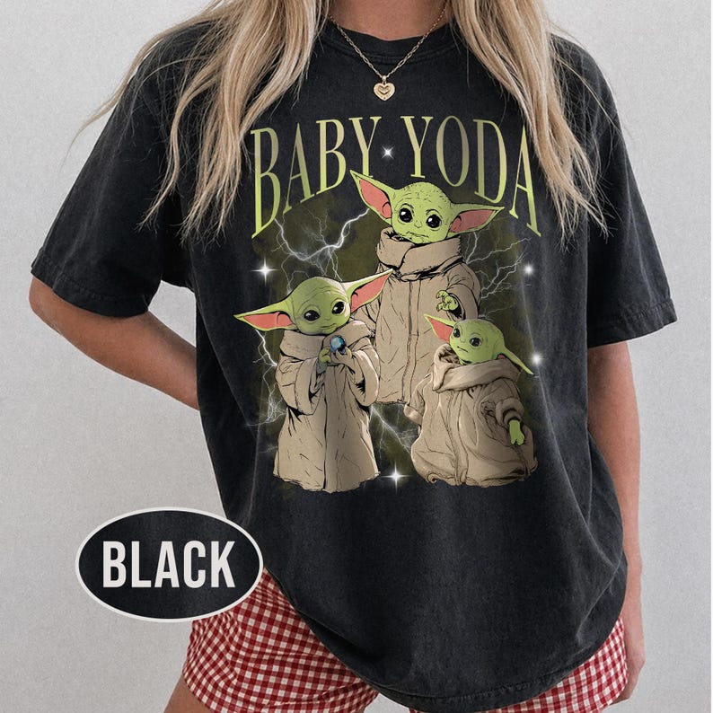 May include: A black short-sleeve t-shirt featuring a graphic print of three green creatures in tan robes with the text BABY YODA in yellow arched lettering above them. The design includes white lightning bolts and small star accents on the front.