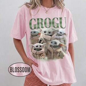 May include: A light pink t-shirt featuring the word "GROGU" in green block letters above a graphic of Grogu, also known as Baby Yoda, from the Star Wars series. The graphic shows Grogu in various poses. The shirt has short sleeves.
