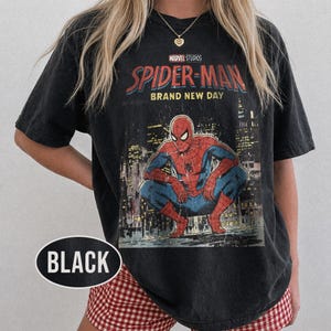 Może przedstawiać: Czarny t-shirt z wytartą grafiką Spider-Mana w kucającej pozie na tle miasta. Koszulka zawiera napis "MARVEL STUDIOS SPIDER-MAN BRAND NEW DAY".