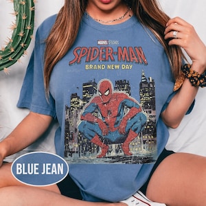 Może przedstawiać: Niebieski t-shirt z kr&oacute;tkim rękawem z grafiką Spider-Mana. Grafika zawiera tekst "Marvel Studios Spider-Man Brand New Day" oraz ilustrację Spider-Mana w miejskim otoczeniu. Koszulka jest w kolorze jasnoniebieskim.