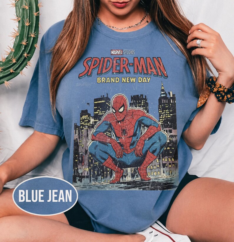 Może przedstawiać: Niebieski t-shirt z kr&oacute;tkim rękawem z grafiką Spider-Mana. Grafika zawiera tekst "Marvel Studios Spider-Man Brand New Day" oraz ilustrację Spider-Mana w miejskim otoczeniu. Koszulka jest w kolorze jasnoniebieskim.