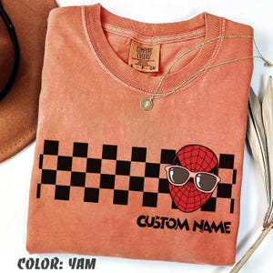May include: A coral-colored t-shirt with a black and white checkered pattern and a graphic of a red spider-like character wearing sunglasses. The text "CUSTOM NAME" is below the graphic. A gold necklace is draped on the shirt.