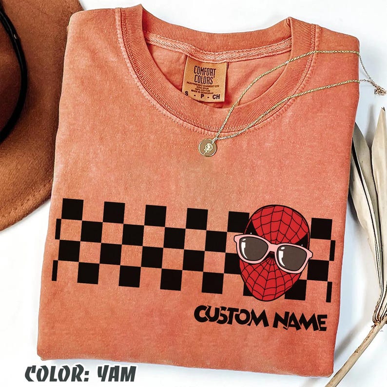 May include: A coral-colored t-shirt with a black and white checkered pattern and a graphic of a red spider-like character wearing sunglasses. The text "CUSTOM NAME" is below the graphic. A gold necklace is draped on the shirt.