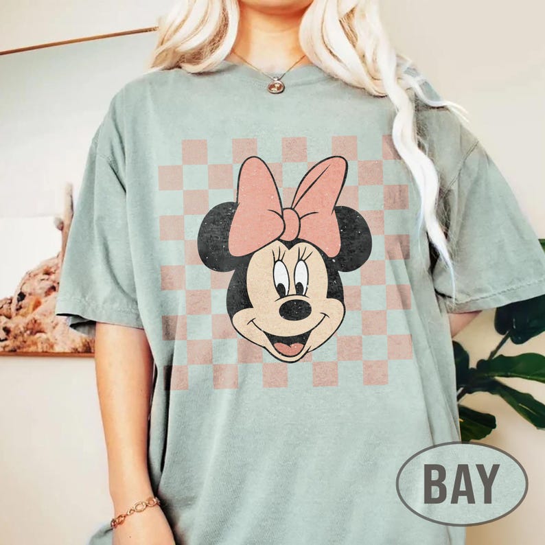 May include: A light green t-shirt featuring a Minnie Mouse graphic. The graphic shows Minnie's face with a pink bow and is set against a pink and white checkered background. The word "BAY" is in a white oval on the bottom right.
