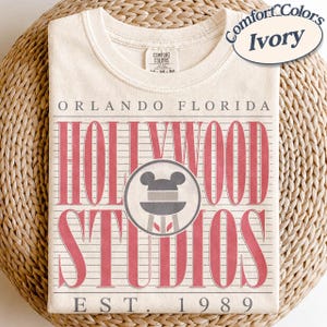 May include: Ivory-coloured t-shirt with a crew neck. The front displays the text "ORLANDO FLORIDA HOLLYWOOD STUDIOS EST. 1989" in red, with a Mickey Mouse icon. The shirt is made by Comfort Colors.