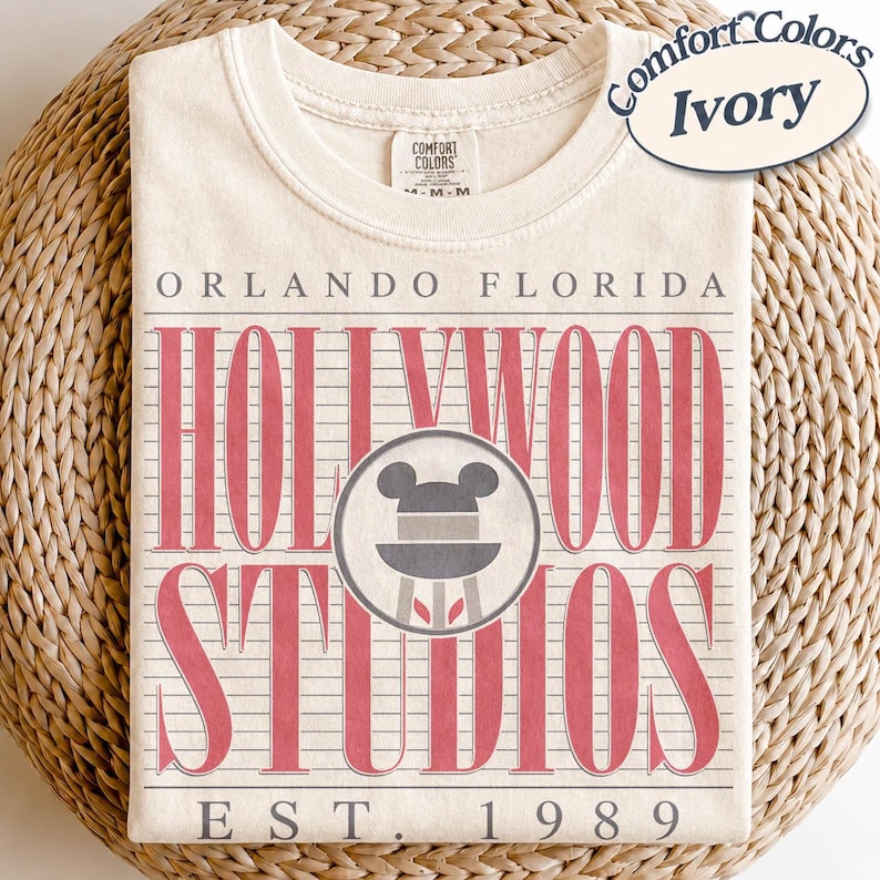 May include: Ivory-coloured t-shirt with a crew neck. The front displays the text "ORLANDO FLORIDA HOLLYWOOD STUDIOS EST. 1989" in red, with a Mickey Mouse icon. The shirt is made by Comfort Colors.