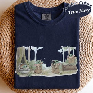 K&ouml;nnte beinhalten: Marineblaues T-Shirt mit einer Grafik von Grogu-Charakteren in einer Waldszene. Das Design umfasst B&auml;ume, einen Baumstamm und Grogu in verschiedenen Posen. Der Text "Comfort Colors True Navy" befindet sich in einem wei&szlig;en Oval auf dem T-Shirt.