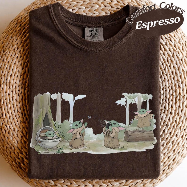 K&ouml;nnte beinhalten: Espressofarbenes T-Shirt mit einer Grafik einer Figur aus einer beliebten Science-Fiction-Serie. Das Design zeigt die Figur in einer Waldkulisse mit B&auml;umen und einem kleinen Frosch. Das Shirt hat das Label der Marke Comfort Colors.