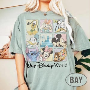 May include: A sage green short-sleeve t-shirt featuring a grid of nine squares with illustrations of Disney characters including Pluto, Daisy Duck, Goofy, Donald Duck, a castle, Mickey Mouse, Stitch, Minnie Mouse, and Chip and Dale, with the text Walt Disney World.