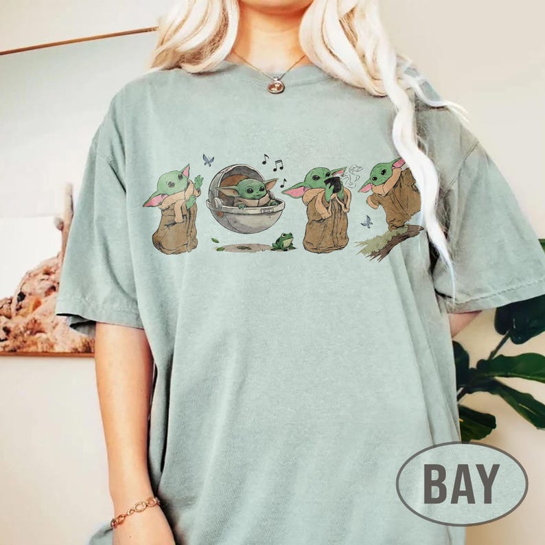 May include: A pale green t-shirt with a graphic of a character from a popular science fiction series. The graphic shows the character in different poses, with musical notes and a frog. The word "BAY" is printed on the bottom right.