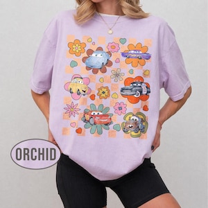 May include: A lilac t-shirt with a retro design featuring cartoon cars and flowers. The cars are in various colours, set against a checkered and floral background. The word "ORCHID" is printed on the shirt.