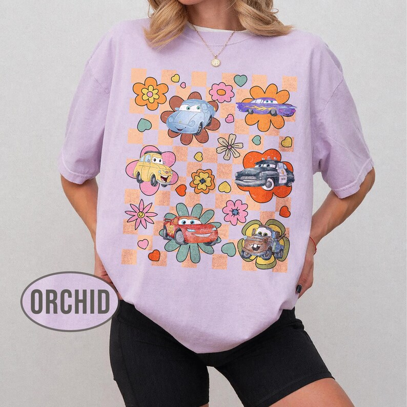 May include: A lilac t-shirt with a retro design featuring cartoon cars and flowers. The cars are in various colours, set against a checkered and floral background. The word "ORCHID" is printed on the shirt.