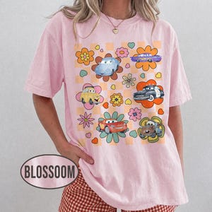 May include: Pale pink t-shirt with a colourful graphic of cartoon cars set within flower designs on a chequered background. The shirt has short sleeves and a relaxed fit. The word "BLOSSOM" is printed on the bottom left of the shirt.
