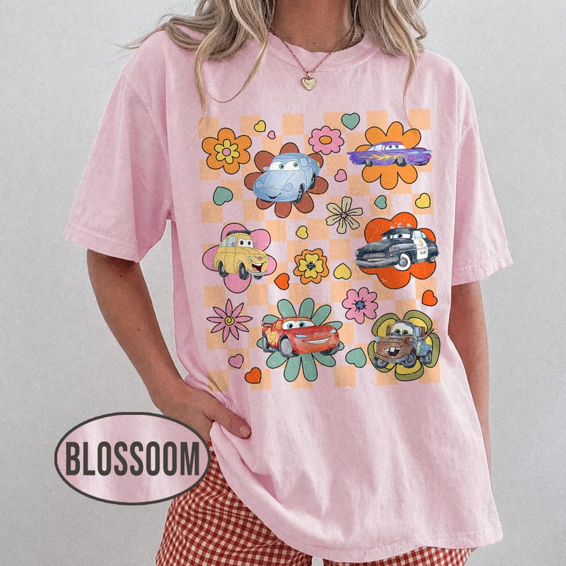 May include: Pale pink t-shirt with a colourful graphic of cartoon cars set within flower designs on a chequered background. The shirt has short sleeves and a relaxed fit. The word "BLOSSOM" is printed on the bottom left of the shirt.