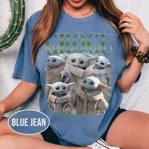 May include: Blue jean-colored t-shirt featuring the word "GROGU" in green lettering above a graphic of Grogu, also known as Baby Yoda, from The Mandalorian. The graphic shows Grogu in various poses. The shirt is a casual style.