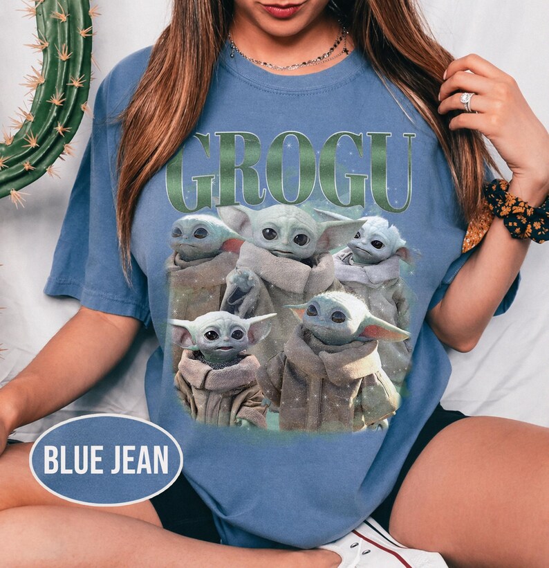 May include: Blue jean-colored t-shirt featuring the word "GROGU" in green lettering above a graphic of Grogu, also known as Baby Yoda, from The Mandalorian. The graphic shows Grogu in various poses. The shirt is a casual style.
