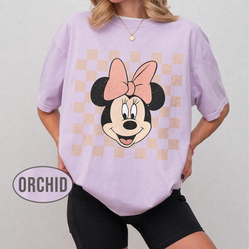 May include: A light purple t-shirt featuring a Minnie Mouse graphic with a pink bow and a checkered background. The word "ORCHID" is printed on the bottom left of the shirt.