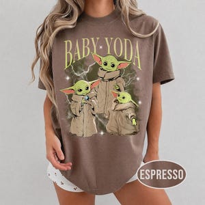 May include: A brown short-sleeve t-shirt featuring a graphic print of three green creatures in tan robes with the text BABY YODA in yellow arched lettering above them. The design includes white lightning bolts and small star accents on the shirt.