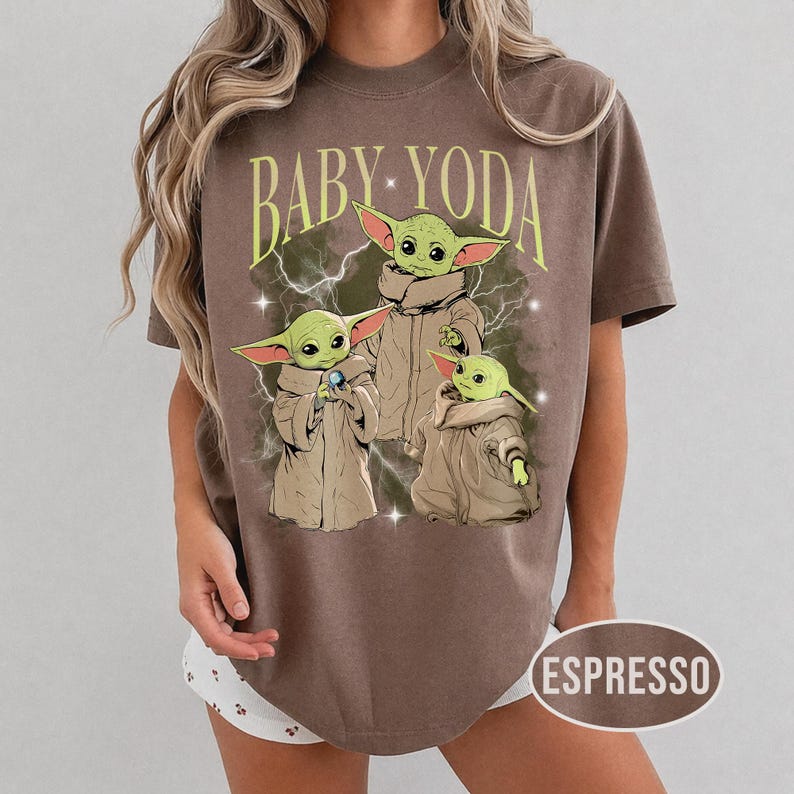 May include: A brown short-sleeve t-shirt featuring a graphic print of three green creatures in tan robes with the text BABY YODA in yellow arched lettering above them. The design includes white lightning bolts and small star accents on the shirt.