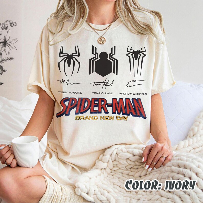 May include: Ivory-colored Spider-Man t-shirt featuring three spider emblems and the names of Tobey Maguire, Tom Holland, and Andrew Garfield. The text "SPIDER-MAN BRAND NEW DAY" is printed in red and blue. The shirt is a relaxed fit.