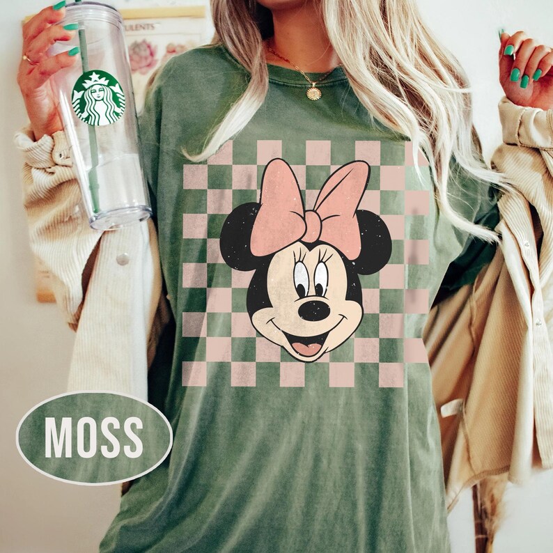 May include: Moss green t-shirt featuring a Minnie Mouse graphic with a pink bow and a checkered background in pink and white. The shirt has a relaxed fit. The word "MOSS" is displayed in a white oval.