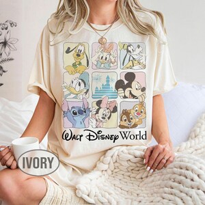 May include: An ivory t-shirt featuring a grid of nine squares, each containing a different Disney character or the castle, with the text Walt Disney World printed in black below the grid. The characters include Pluto, Daisy, Goofy, Donald, Mickey, Stitch, Minnie, Chip, and Dale.