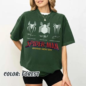 May include: Forest green Spider-Man t-shirt featuring three spider symbols and the names of the actors who played Spider-Man: Tobey Maguire, Tom Holland, and Andrew Garfield. The text "SPIDER-MAN BRAND NEW DAY" is printed in red and blue.