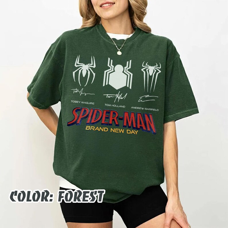 May include: Forest green Spider-Man t-shirt featuring three spider symbols and the names of the actors who played Spider-Man: Tobey Maguire, Tom Holland, and Andrew Garfield. The text "SPIDER-MAN BRAND NEW DAY" is printed in red and blue.