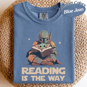 May include: Blue t-shirt featuring a graphic of a character in armour reading a book with a small green creature. The text "Reading is the Way" is printed below the illustration. The shirt is a Comfort Colors brand.