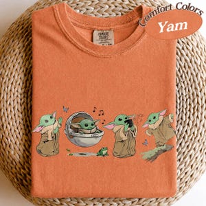 May include: Orange Comfort Colors t-shirt featuring a cartoon character from a popular science fiction series. The design shows the character in different poses, with a frog, butterflies, and musical notes. The shirt has a crew neck.