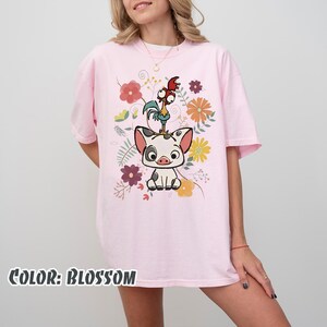 Puede incluir: Camiseta rosa claro con un dise&ntilde;o de cerdo, gallo y flores de dibujos animados. El texto "COLOR: BLOSSOM" est&aacute; en la parte inferior. La camiseta tiene mangas cortas y un corte holgado, ideal para un look casual.