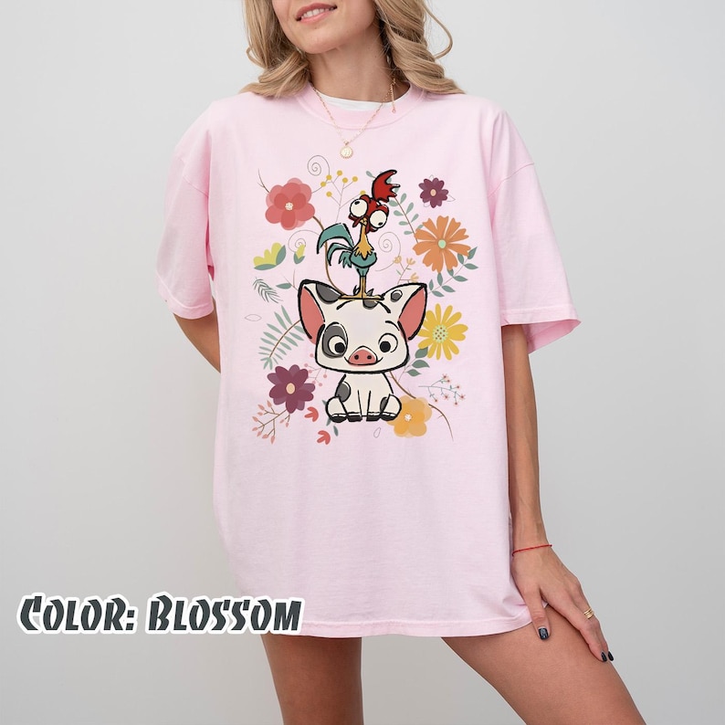 Puede incluir: Camiseta rosa claro con un dise&ntilde;o de cerdo, gallo y flores de dibujos animados. El texto "COLOR: BLOSSOM" est&aacute; en la parte inferior. La camiseta tiene mangas cortas y un corte holgado, ideal para un look casual.