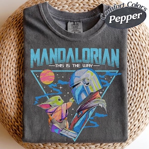 May include: Dark grey t-shirt with a graphic of The Mandalorian and Grogu. The text "MANDALORIAN" and "THIS IS THE WAY" are in blue. The design includes a triangle, a planet, and stars. The shirt is labelled "Comfort Colors Pepper".