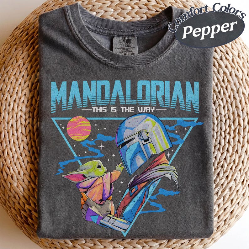May include: Dark grey t-shirt with a graphic of The Mandalorian and Grogu. The text "MANDALORIAN" and "THIS IS THE WAY" are in blue. The design includes a triangle, a planet, and stars. The shirt is labelled "Comfort Colors Pepper".