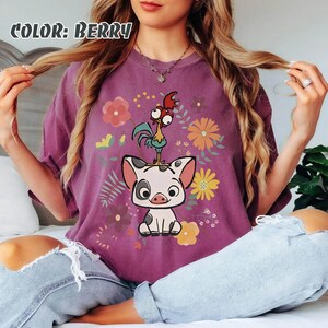 Puede incluir: Camiseta de color bayas con un dise&ntilde;o de cerdo y gallo de dibujos animados, rodeado de flores. El texto "COLOR: BERRY" est&aacute; en la parte superior. El gr&aacute;fico est&aacute; centrado en la parte delantera de la camiseta.