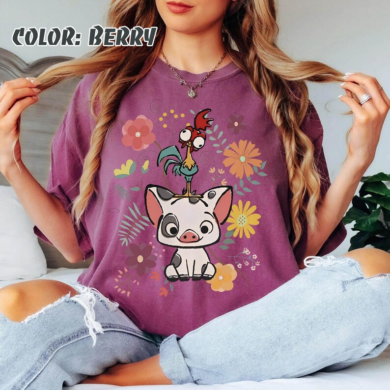 Puede incluir: Camiseta de color bayas con un dise&ntilde;o de cerdo y gallo de dibujos animados, rodeado de flores. El texto "COLOR: BERRY" est&aacute; en la parte superior. El gr&aacute;fico est&aacute; centrado en la parte delantera de la camiseta.