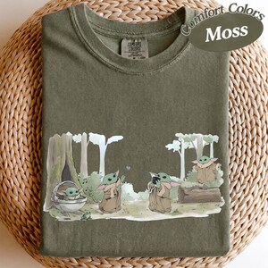 K&ouml;nnte beinhalten: Moosgr&uuml;nes T-Shirt mit einer Grafik von Grogu aus The Mandalorian. Das Design zeigt Grogu in verschiedenen Posen in einer Waldkulisse. Auf dem Shirt stehen die Worte "Comfort Colors" und "Moss".