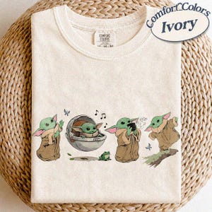 May include: Ivory Comfort Colors t-shirt with a graphic of a green alien character in different poses. The design includes a frog, butterflies, and musical notes. The t-shirt has a crew neck and short sleeves.