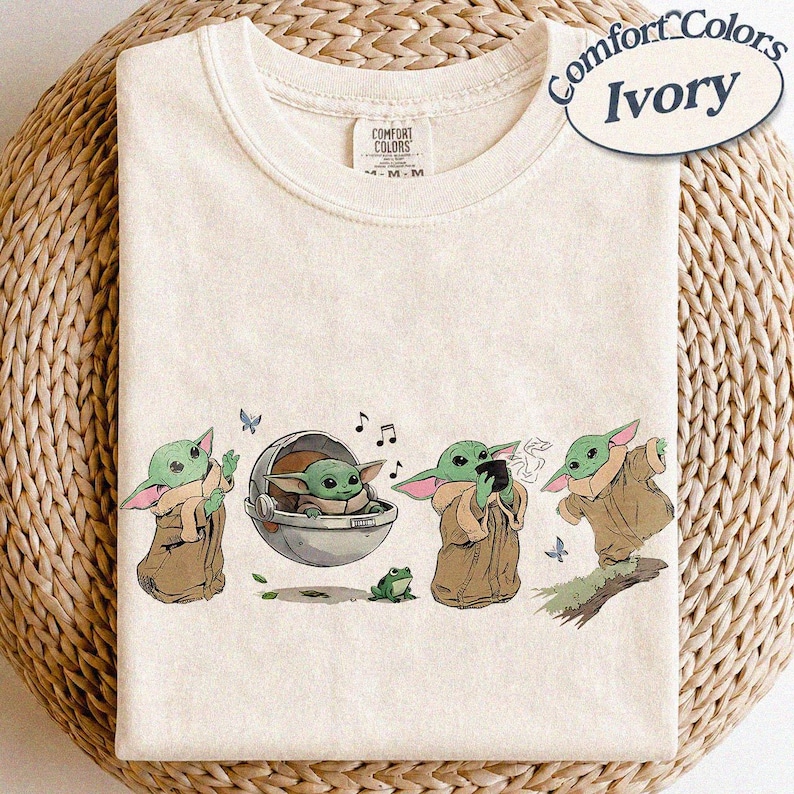 May include: Ivory Comfort Colors t-shirt with a graphic of a green alien character in different poses. The design includes a frog, butterflies, and musical notes. The t-shirt has a crew neck and short sleeves.