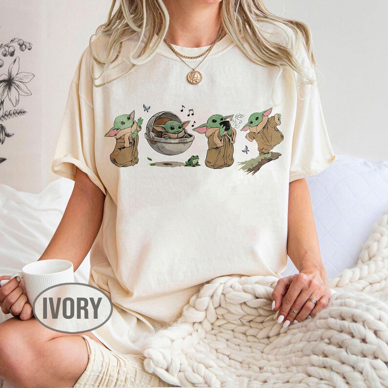 May include: Cream-coloured t-shirt with a graphic of a character from a popular science fiction series. The design shows the character in various poses, with musical notes and a frog. The word "IVORY" is printed on a mug.