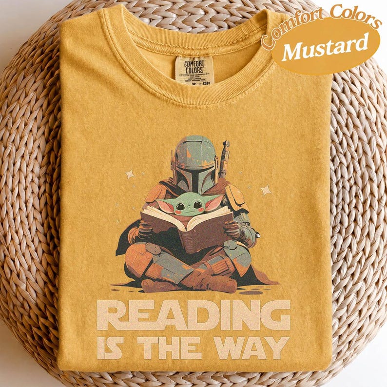 May include: Mustard-coloured t-shirt featuring a graphic of a Mandalorian character reading a book with Grogu. The text "READING IS THE WAY" is printed below the illustration. The shirt is folded on a woven surface.
