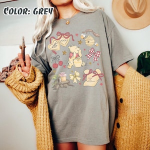 May include: A grey t-shirt with a Winnie the Pooh design. The design includes illustrations of Pooh Bear, honey pots, bows, and the text "Winnie the Pooh". The shirt has short sleeves and a relaxed fit.