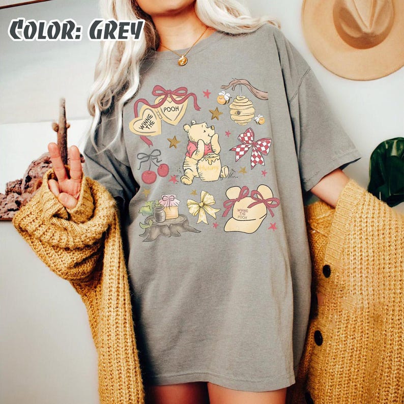 May include: A grey t-shirt with a Winnie the Pooh design. The design includes illustrations of Pooh Bear, honey pots, bows, and the text "Winnie the Pooh". The shirt has short sleeves and a relaxed fit.