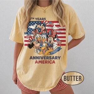 以下が含まれることがあります： バター色のTシャツ。愛国的な衣装を着たディズニーキャラクターのグラフィックが特徴です。デザインにはアメリカ国旗と「250th Years Anniversary America」の文字が含まれています。 ゆったりとしたフィット感のシャツです。