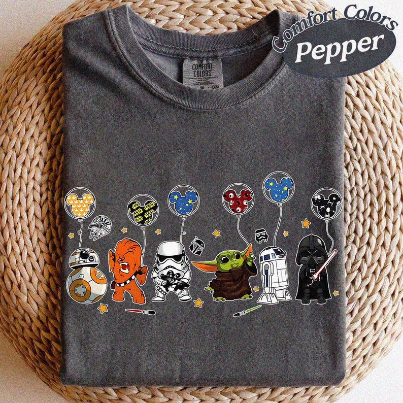 May include: A dark gray t-shirt featuring a line of Star Wars characters, including BB-8, Chewbacca, a Stormtrooper, Grogu, R2-D2, and Darth Vader. Each character is holding a balloon with a Mickey Mouse head design. The shirt has the words "Comfort Colors Pepper" printed on it.