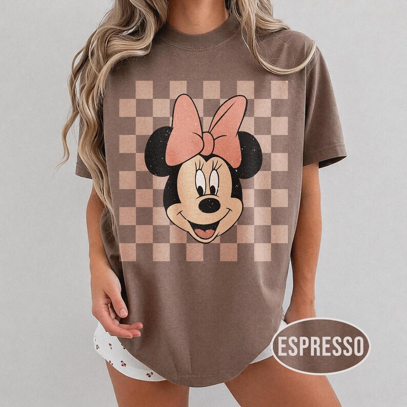 May include: Espresso-colored t-shirt featuring a Minnie Mouse graphic. The design includes a checkered background in shades of brown and a large pink bow. The shirt has a relaxed fit and short sleeves.