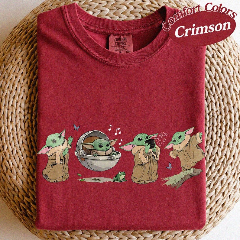 May include: Crimson Comfort Colors t-shirt with a graphic of a green alien character in different poses. The design includes a floating pod, a frog, musical notes, and butterflies. The shirt has a crew neck.
