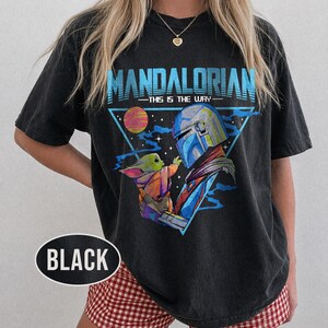 May include: Black t-shirt with a graphic of The Mandalorian and Grogu, and the text "MANDALORIAN - THIS IS THE WAY" in blue. The design includes a planet and a triangular frame. The shirt is a dark colour.